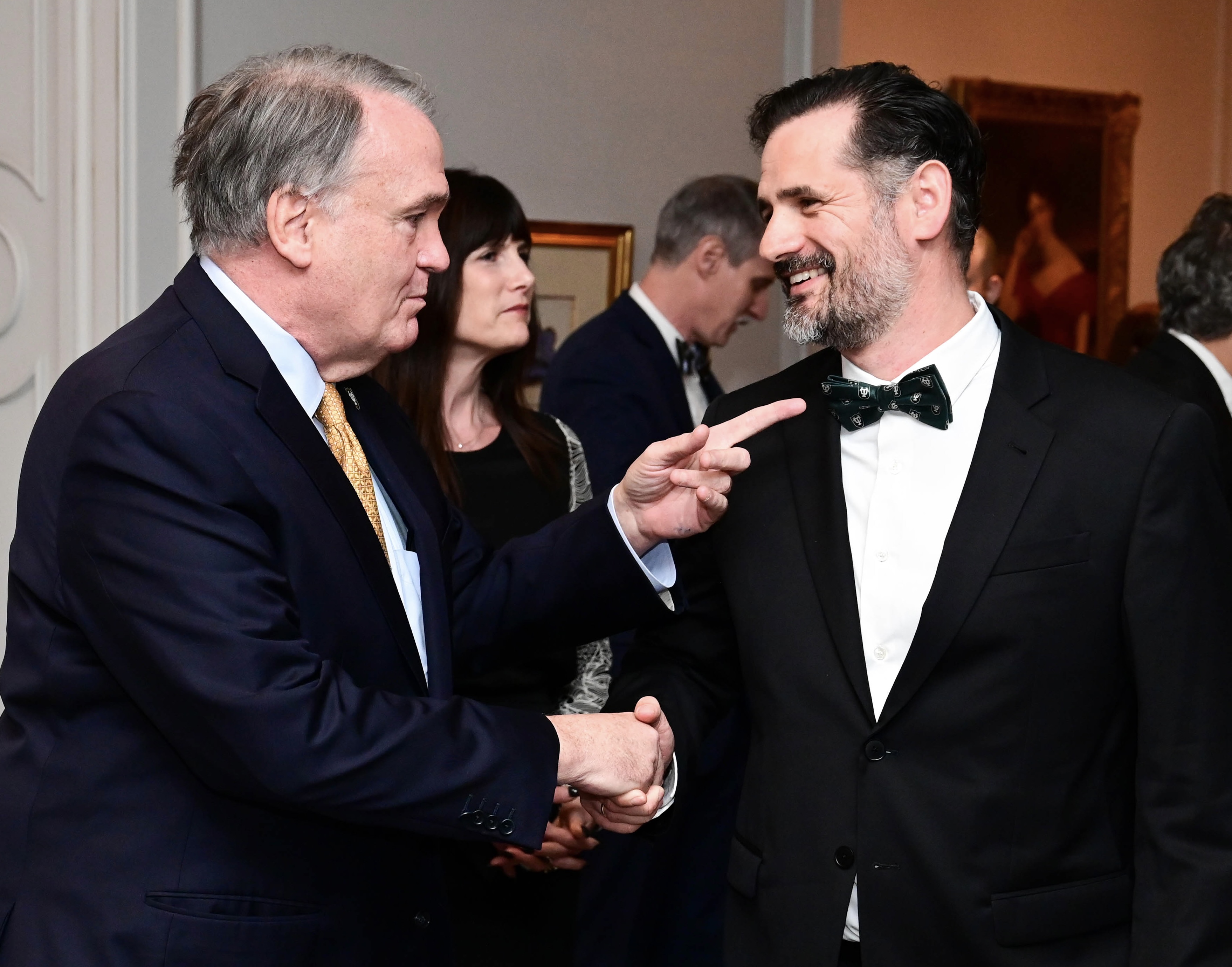 Man in suit shakes hand with smiling man in tuxedo, pointing at his Tulane bowtie.