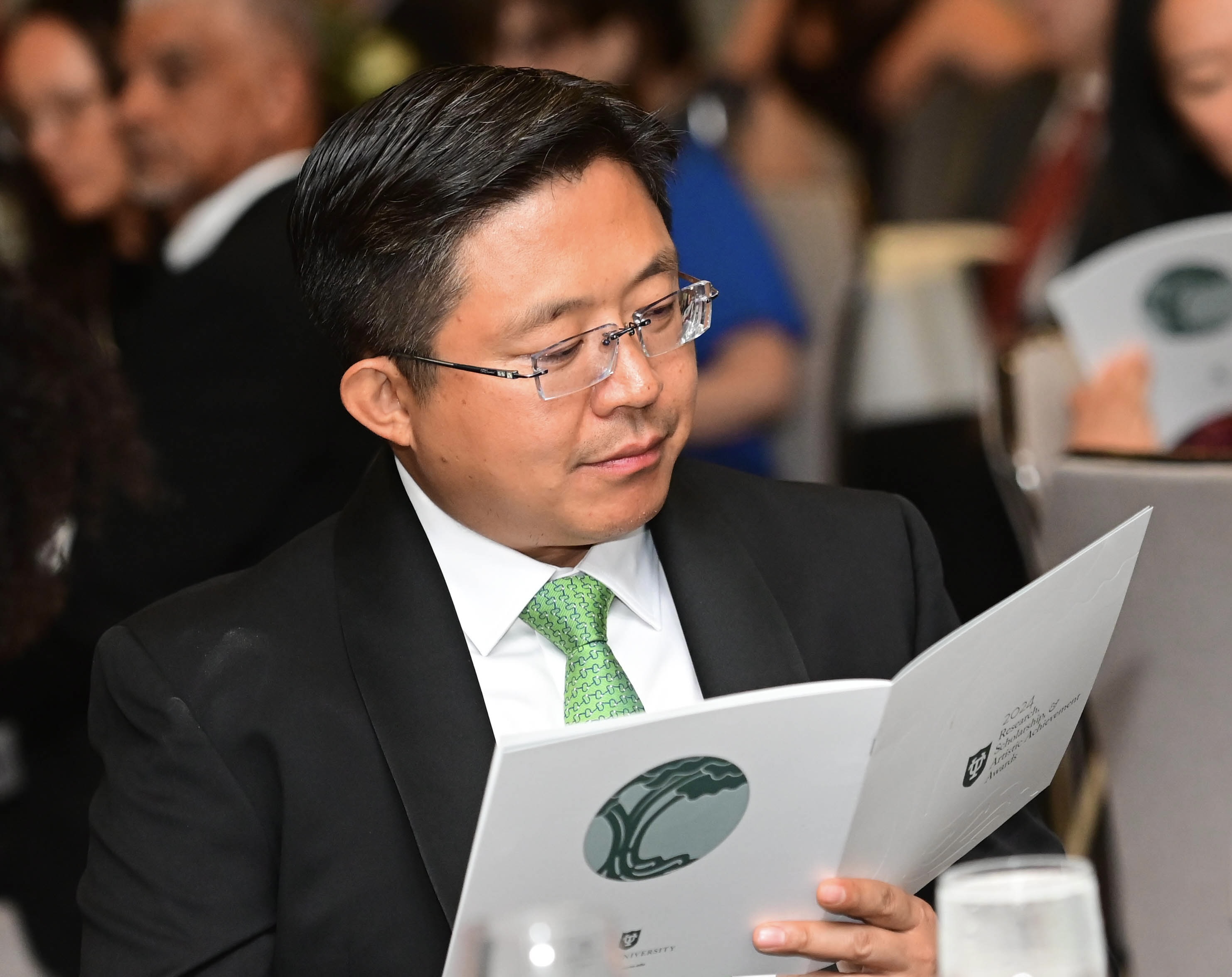 Man in a suit and green tie reads a booklet at a formal event.