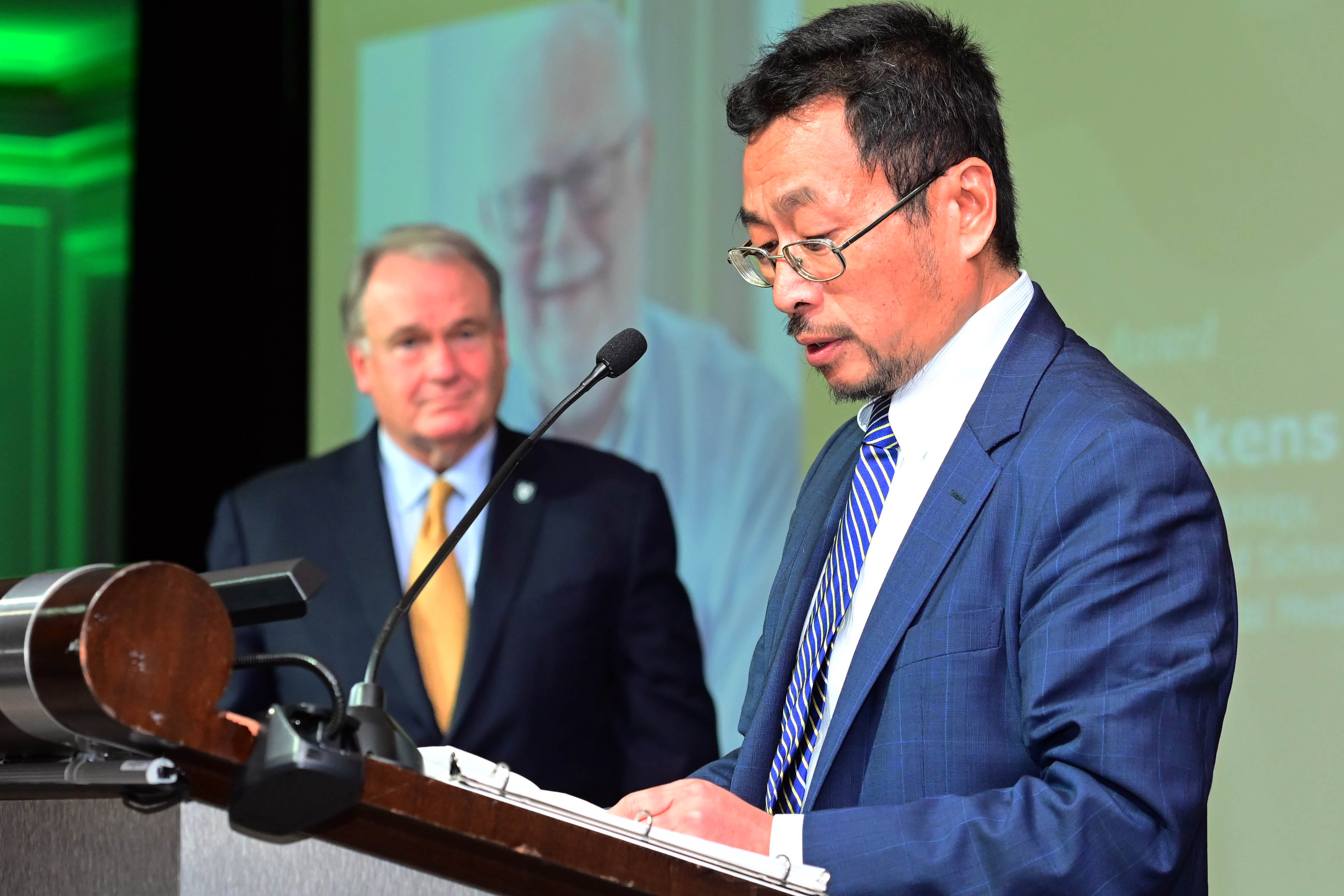 Man in suit speaks at podium, another man in suit stands behind him.