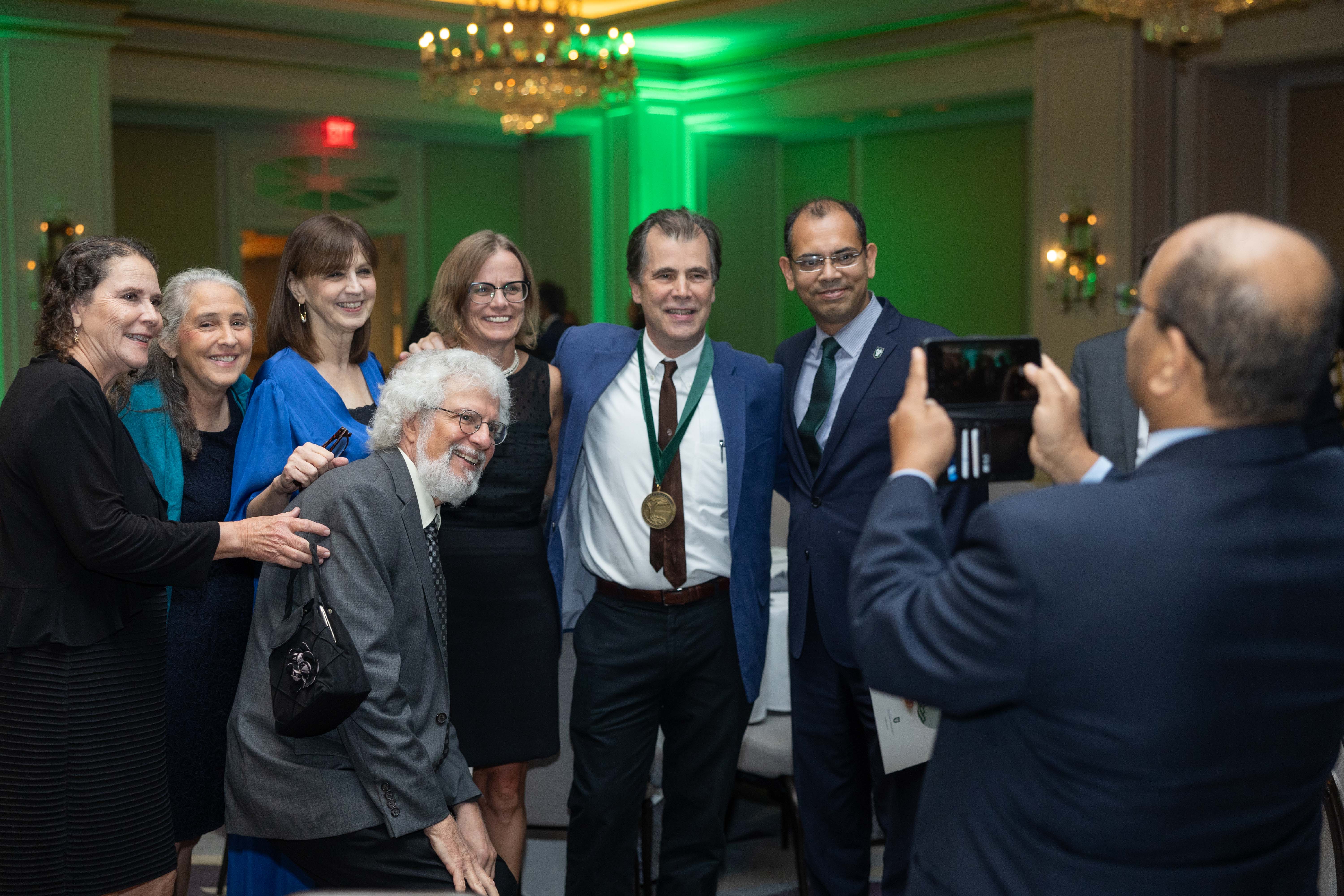 Group of smiling people at a formal event, man with medal, another man taking a photo.