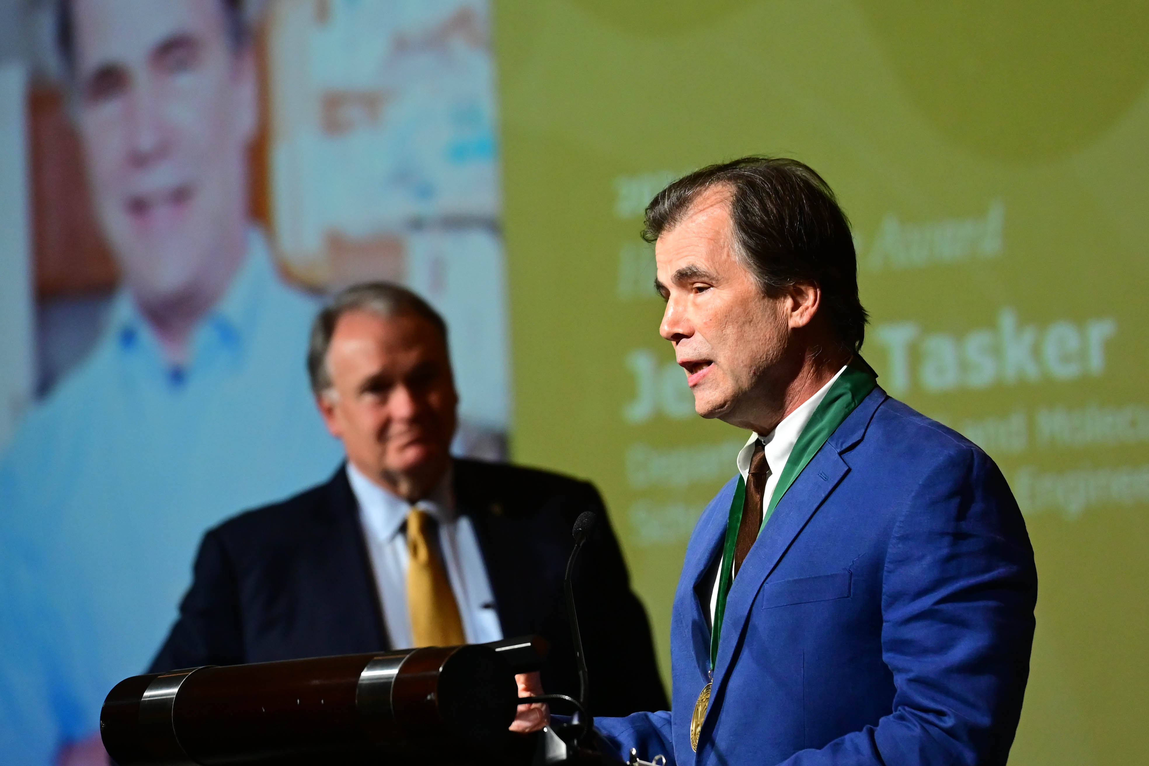 Man in blue suit with medal speaking at a podium; another man and screen in background.