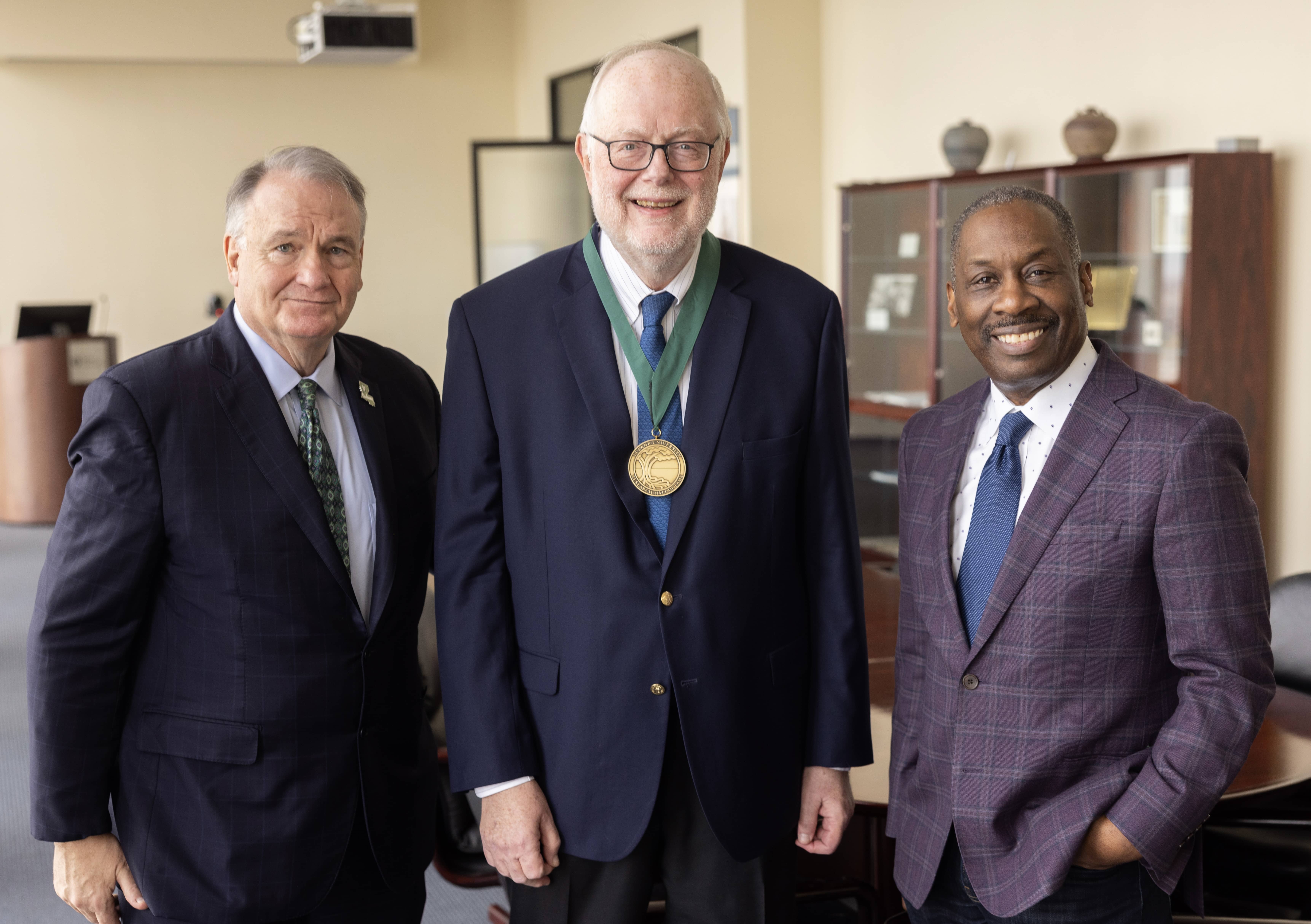 Three smiling men, one with a gold medal, stand in an office.