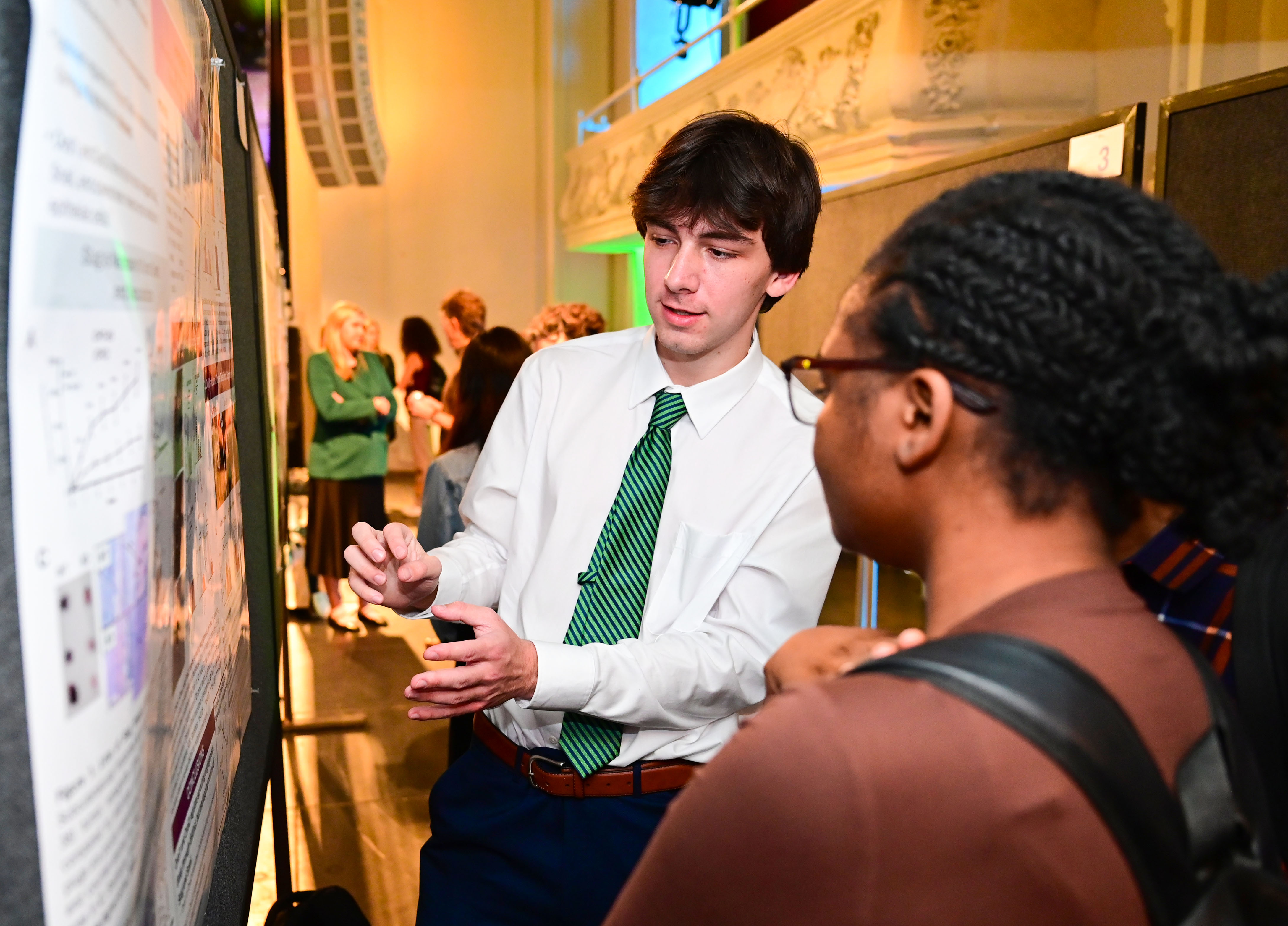 Student in green tie explains research poster to woman with glasses at a conference.