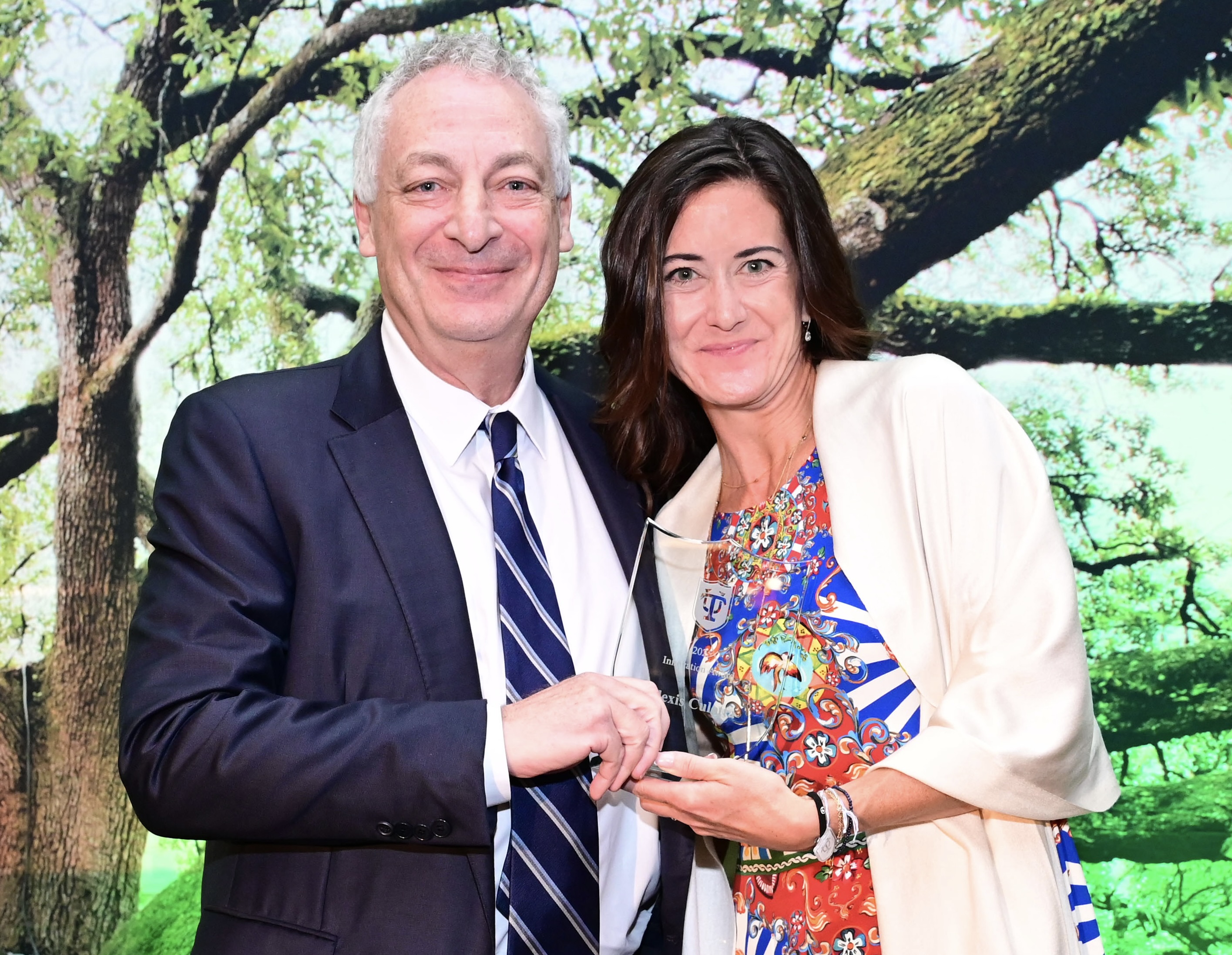 Man in suit and woman in dress smiling, holding a glass award. Green tree backdrop.