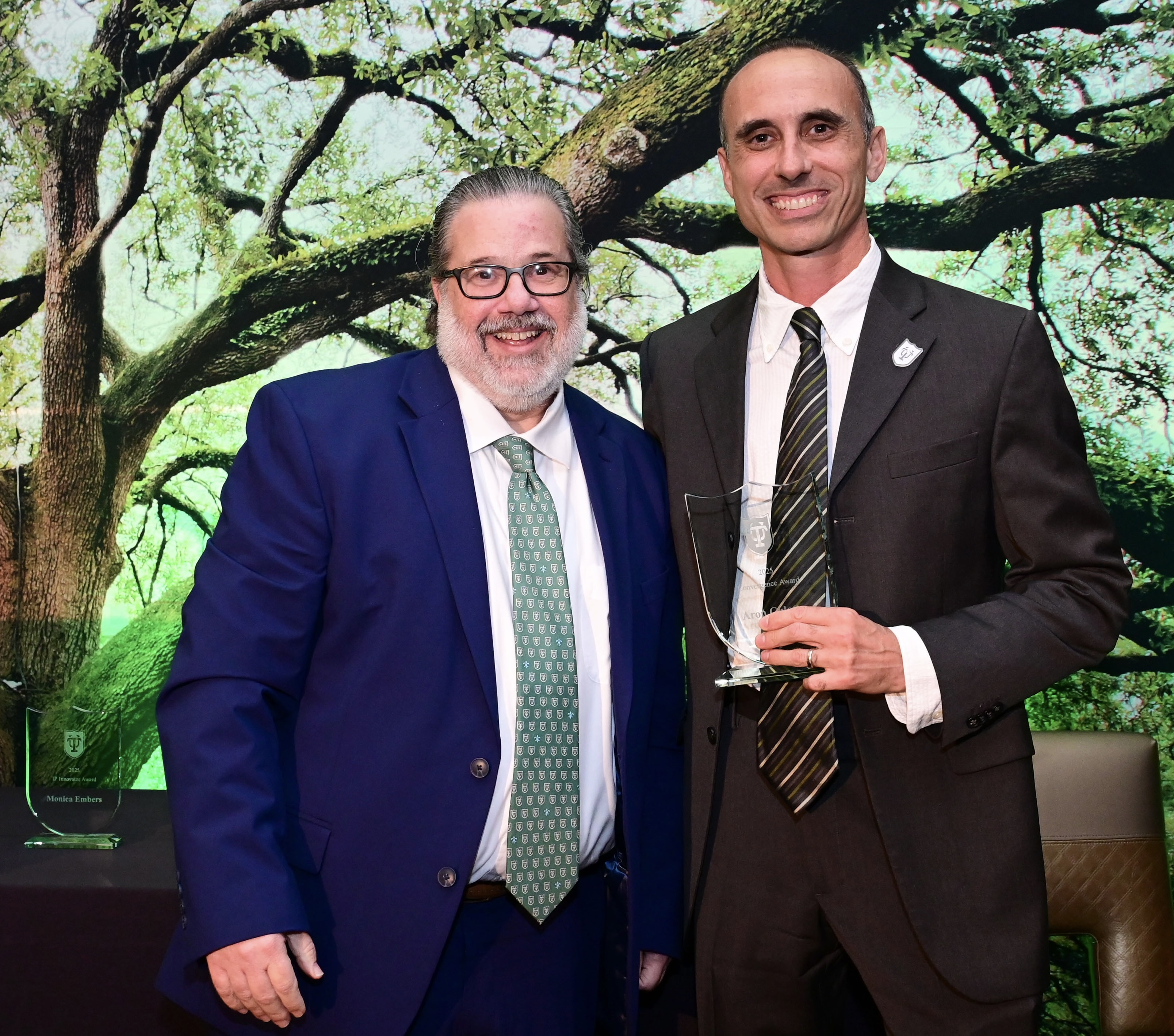 Two smiling men in suits, taller man holding a glass award trophy. Green tree background.