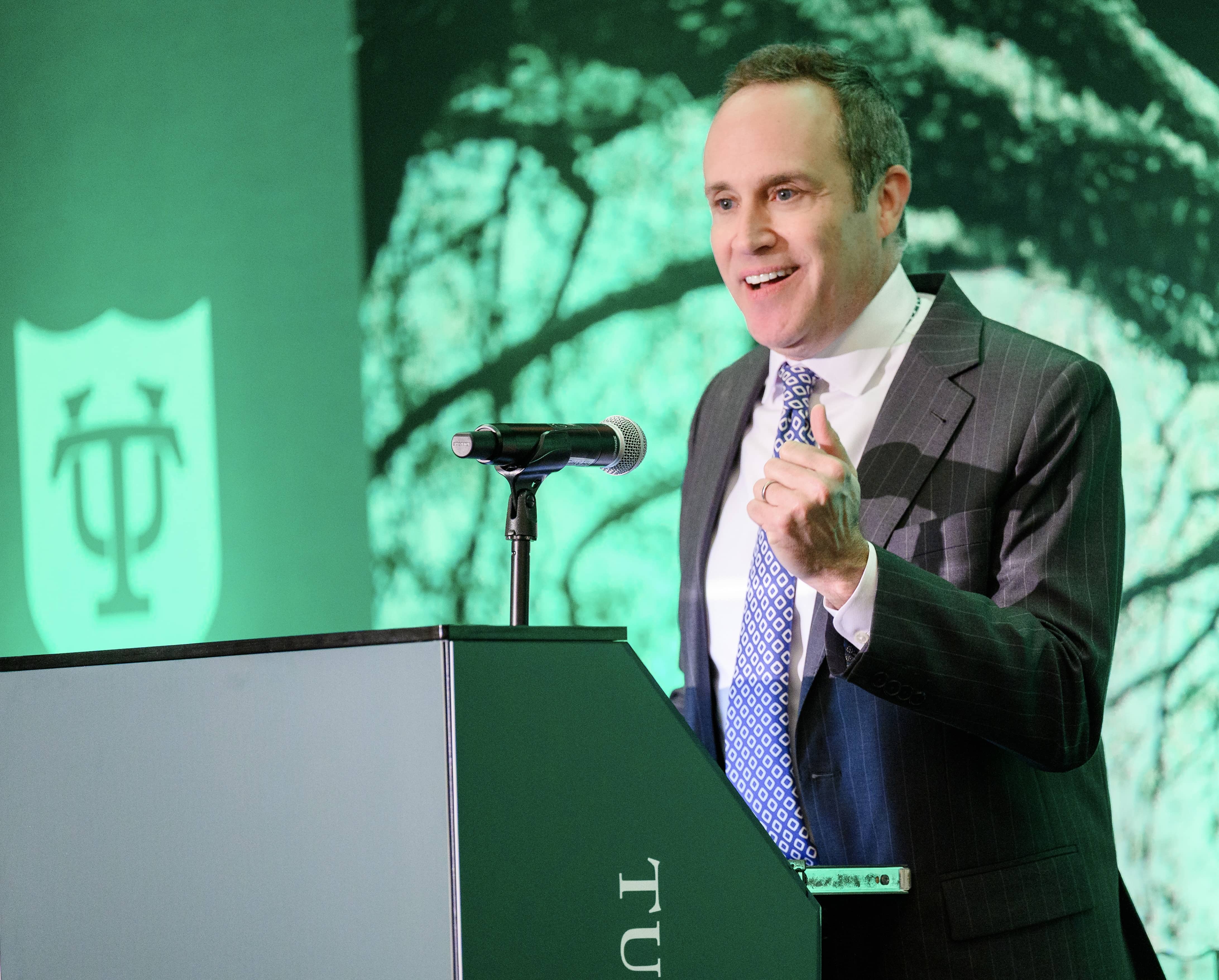 Man in suit speaking at podium with microphone, green background, crest logo. David Bernard, Emcee.