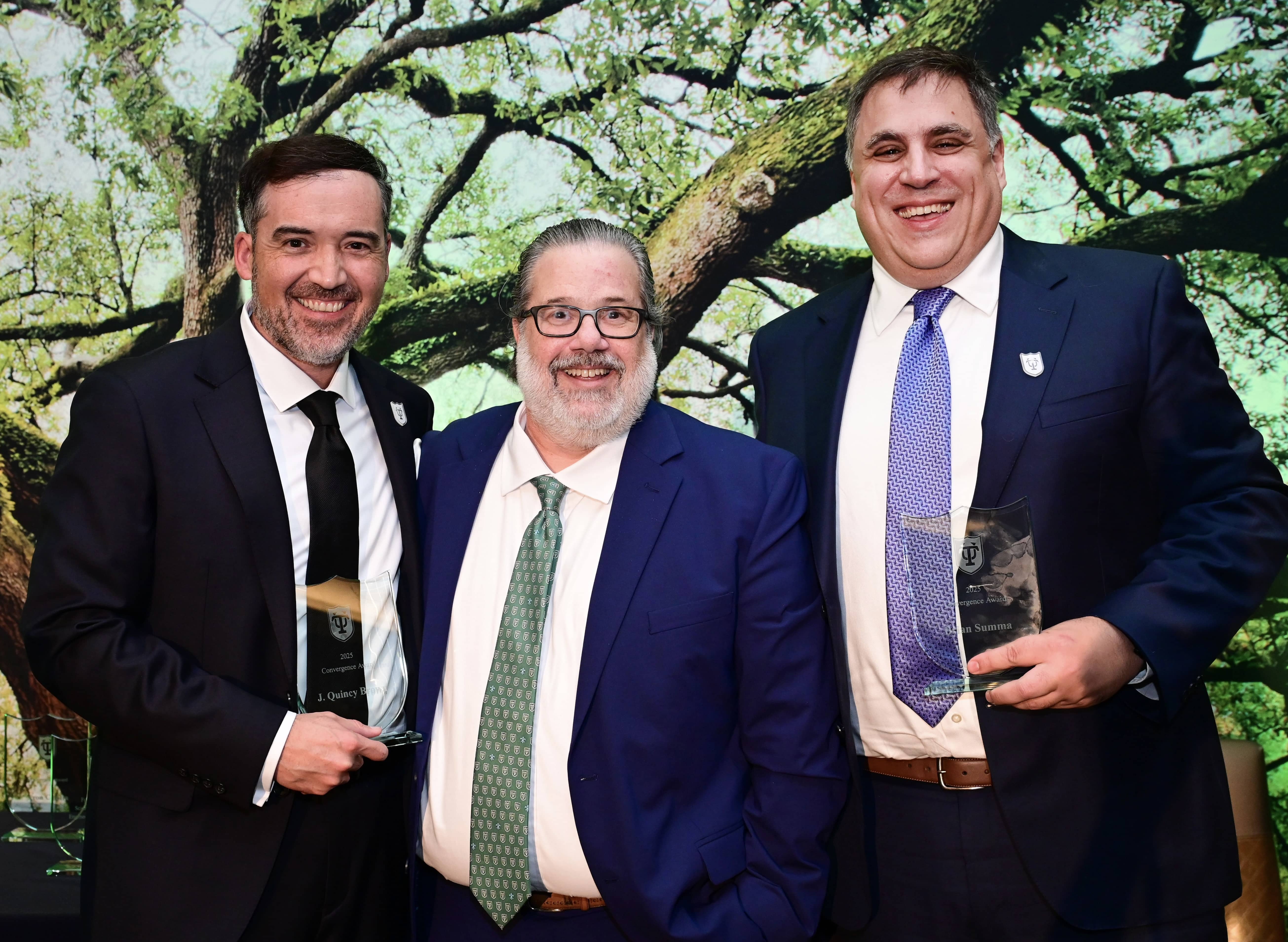 Three smiling men in suits hold awards in front of a green tree backdrop.