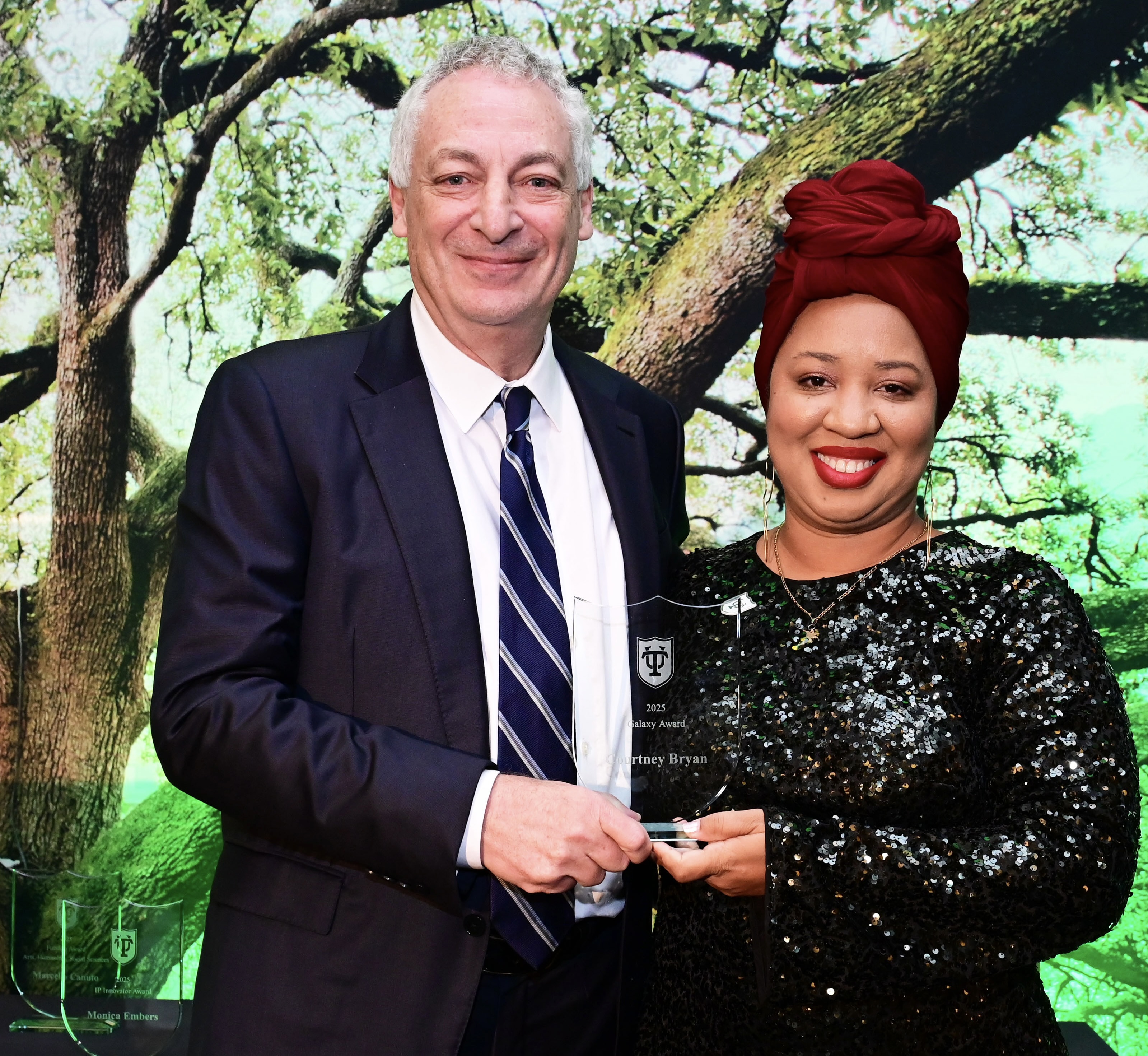 Man in suit and woman in sequined dress, red headwrap, smile while holding an award.