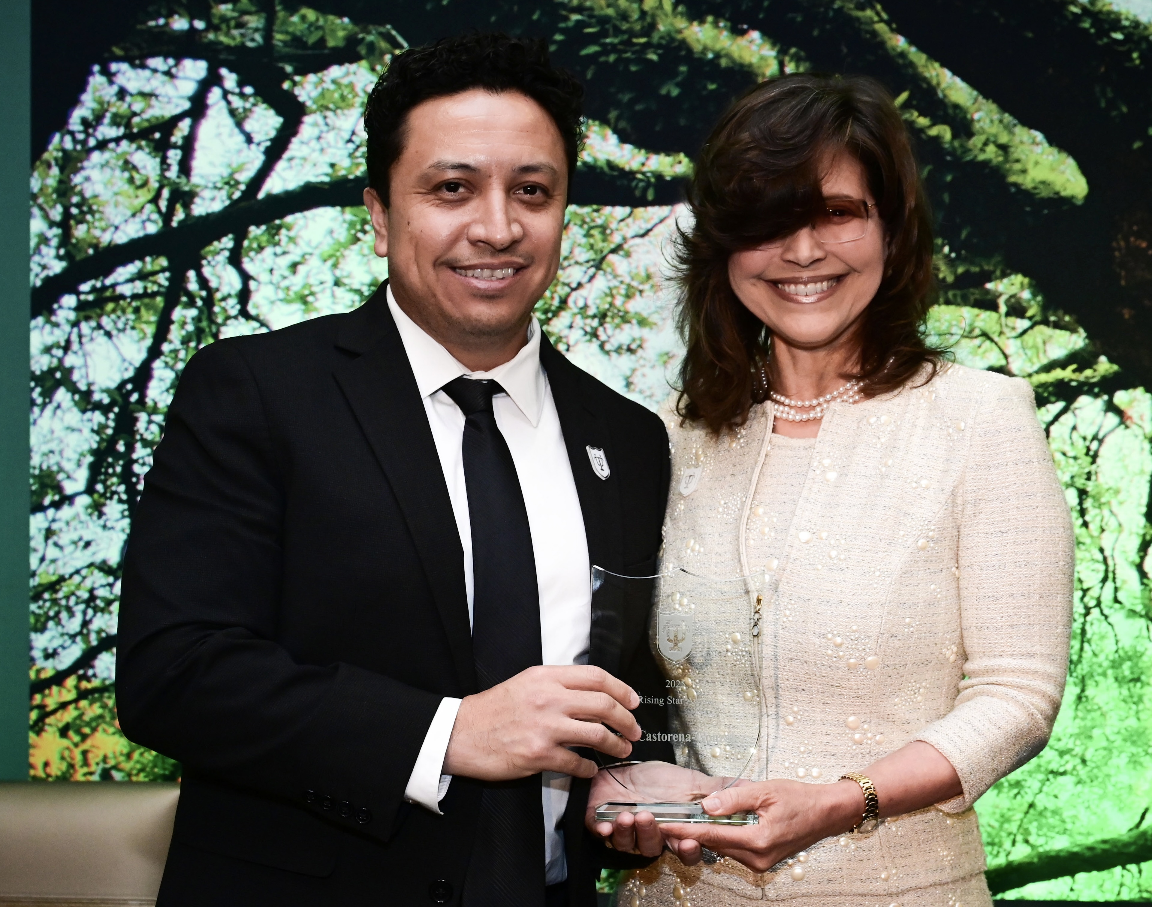 Man in suit and woman in cream dress smile, holding a glass award at a presentation.