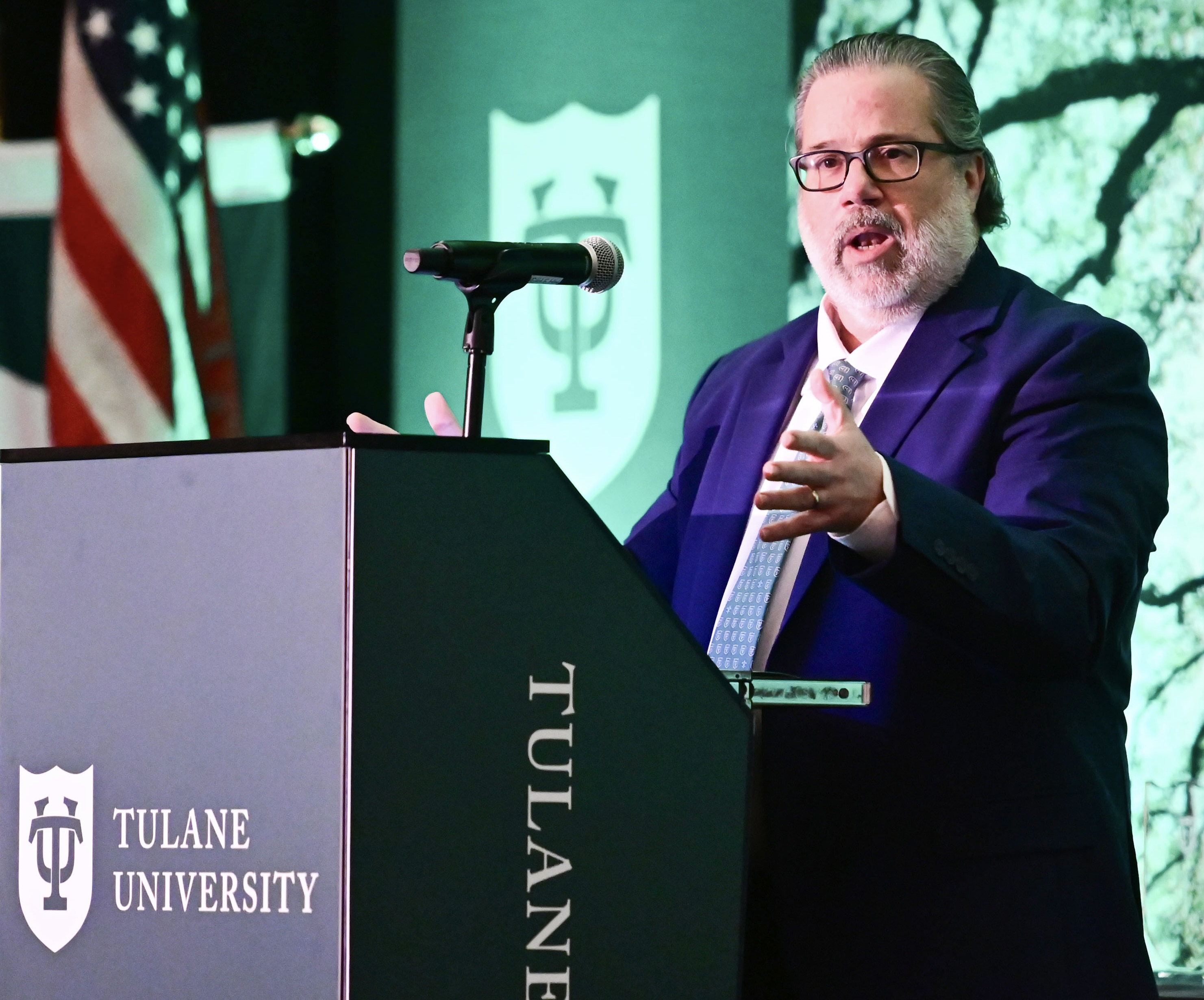 Bearded man in suit and glasses speaks at a Tulane University podium; American flag in background.