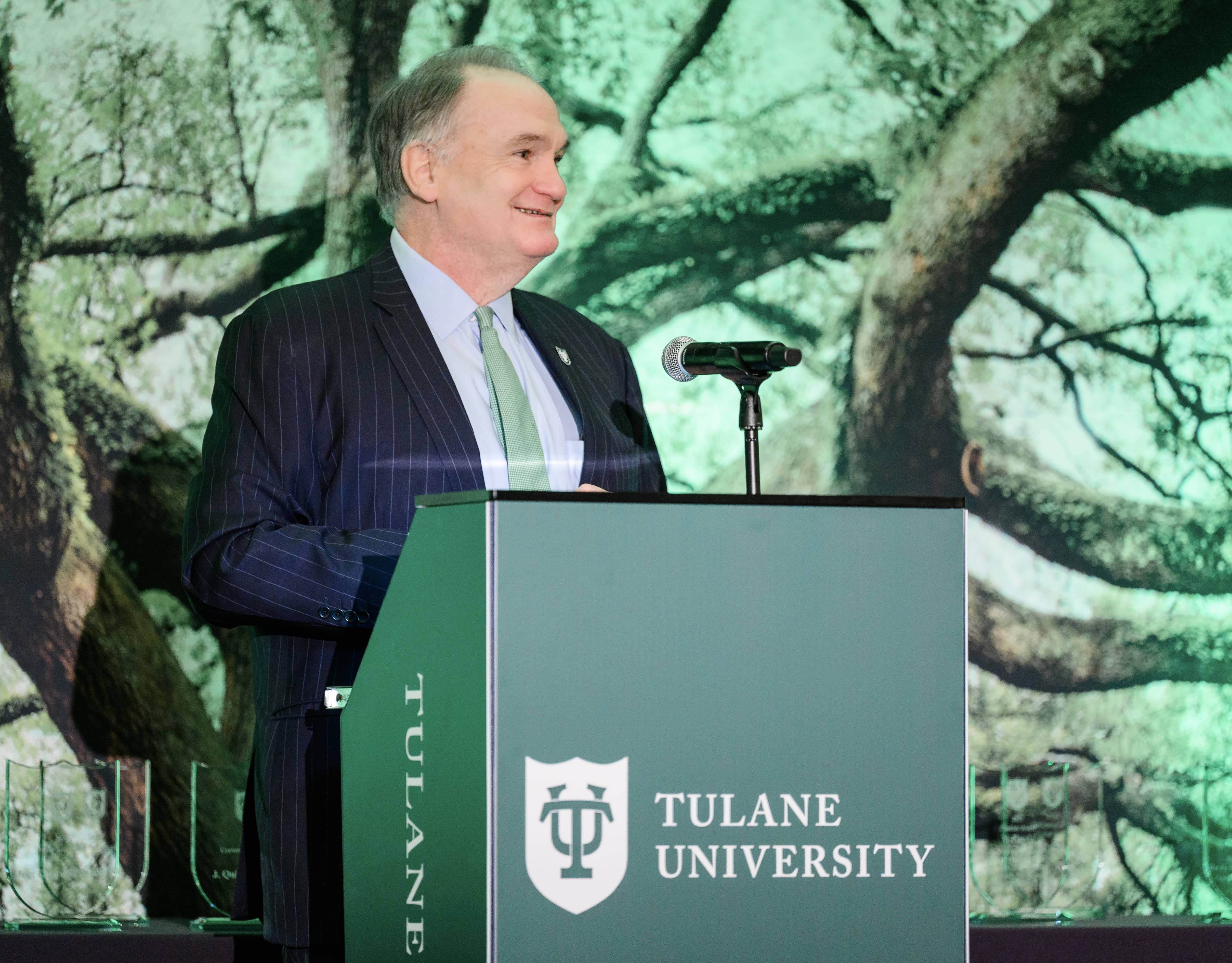 Smiling man in suit speaks at green Tulane University podium with tree-like background.