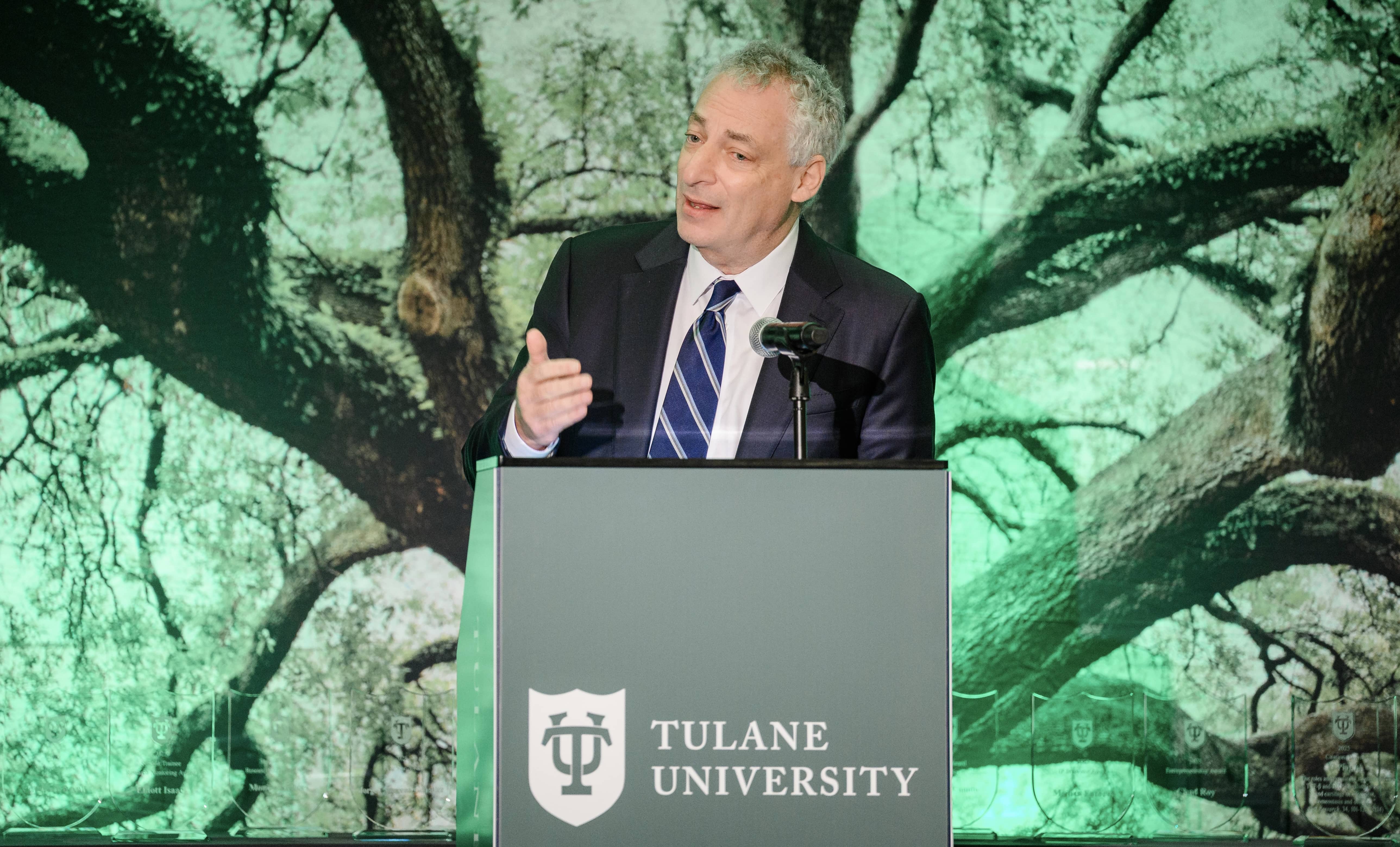 Man in suit speaks at Tulane University podium with green tree background.