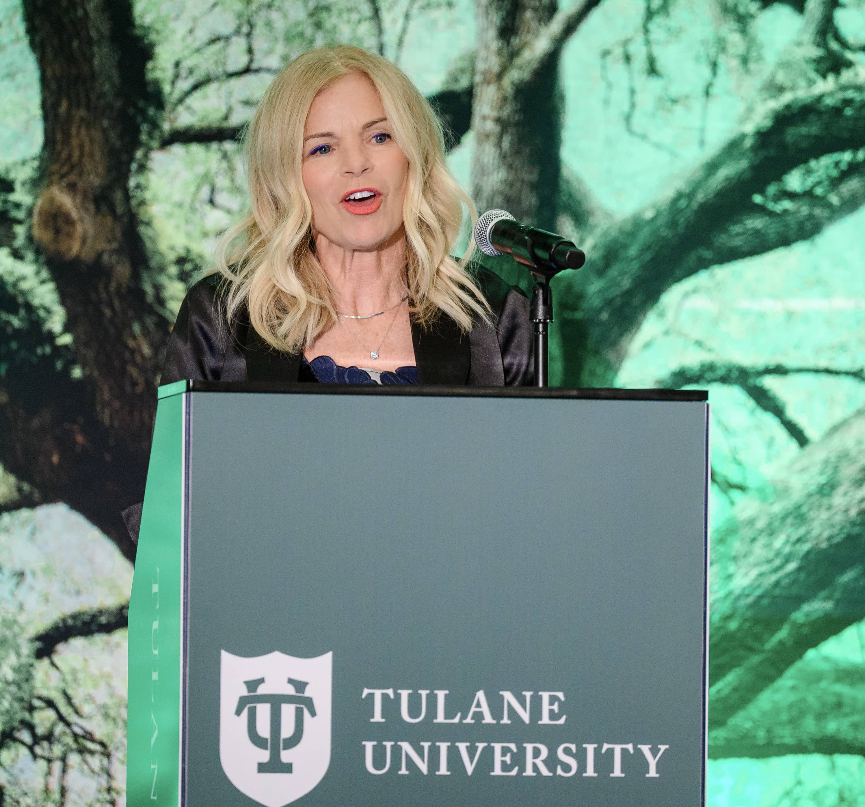 Blonde woman speaking at a Tulane University podium with green tree background.