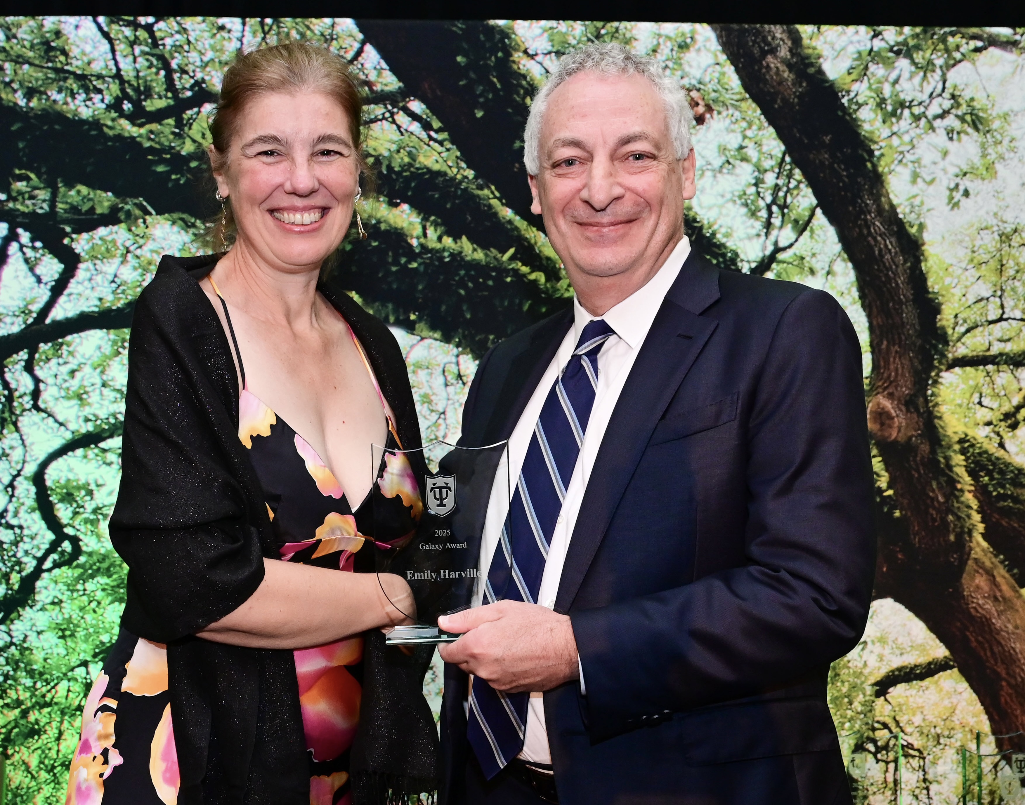 Smiling woman in dress presents a clear award plaque to a man in a suit, green foliage background.
