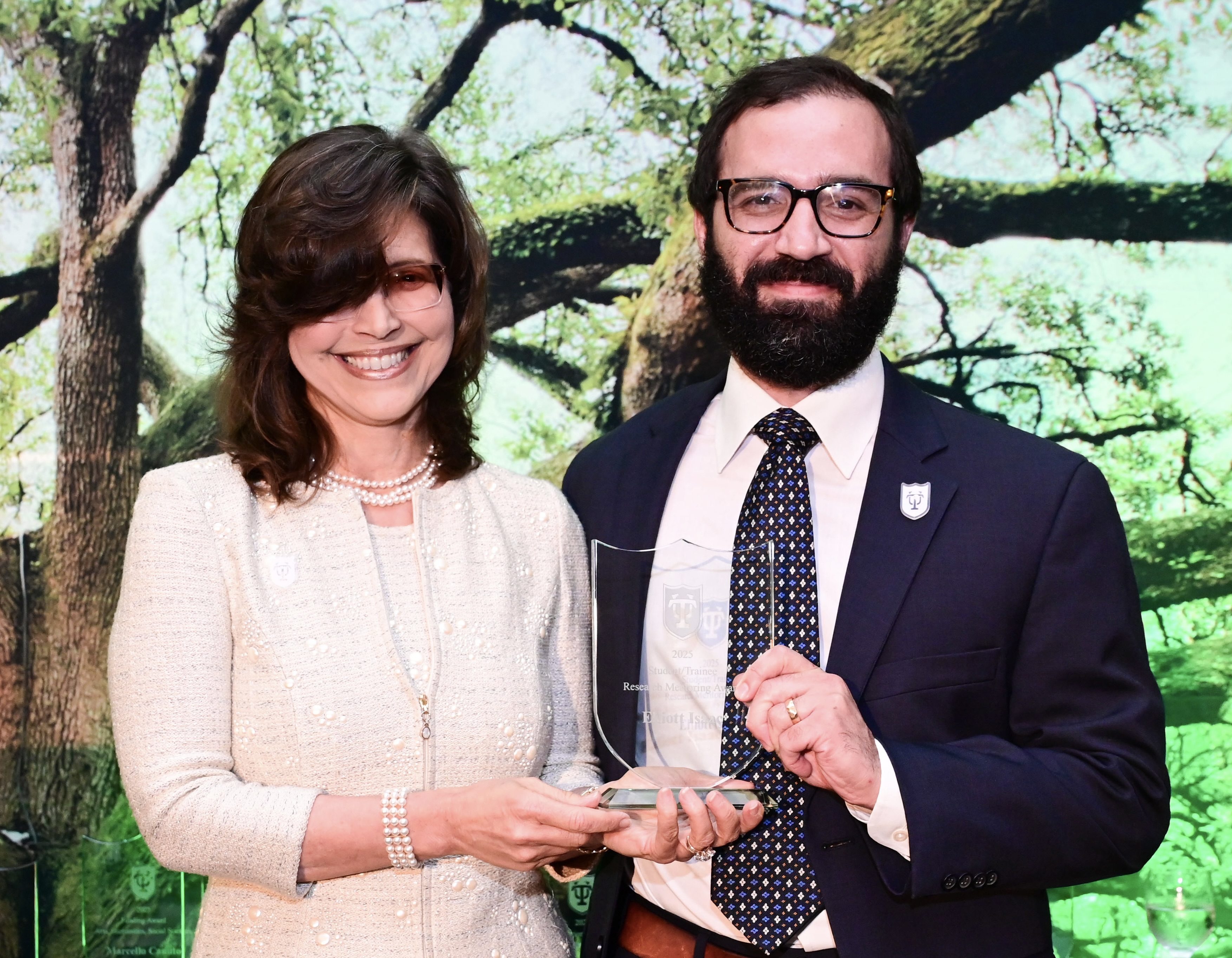 Photograph of a smiling woman and man holding a clear award against a tree backdrop.