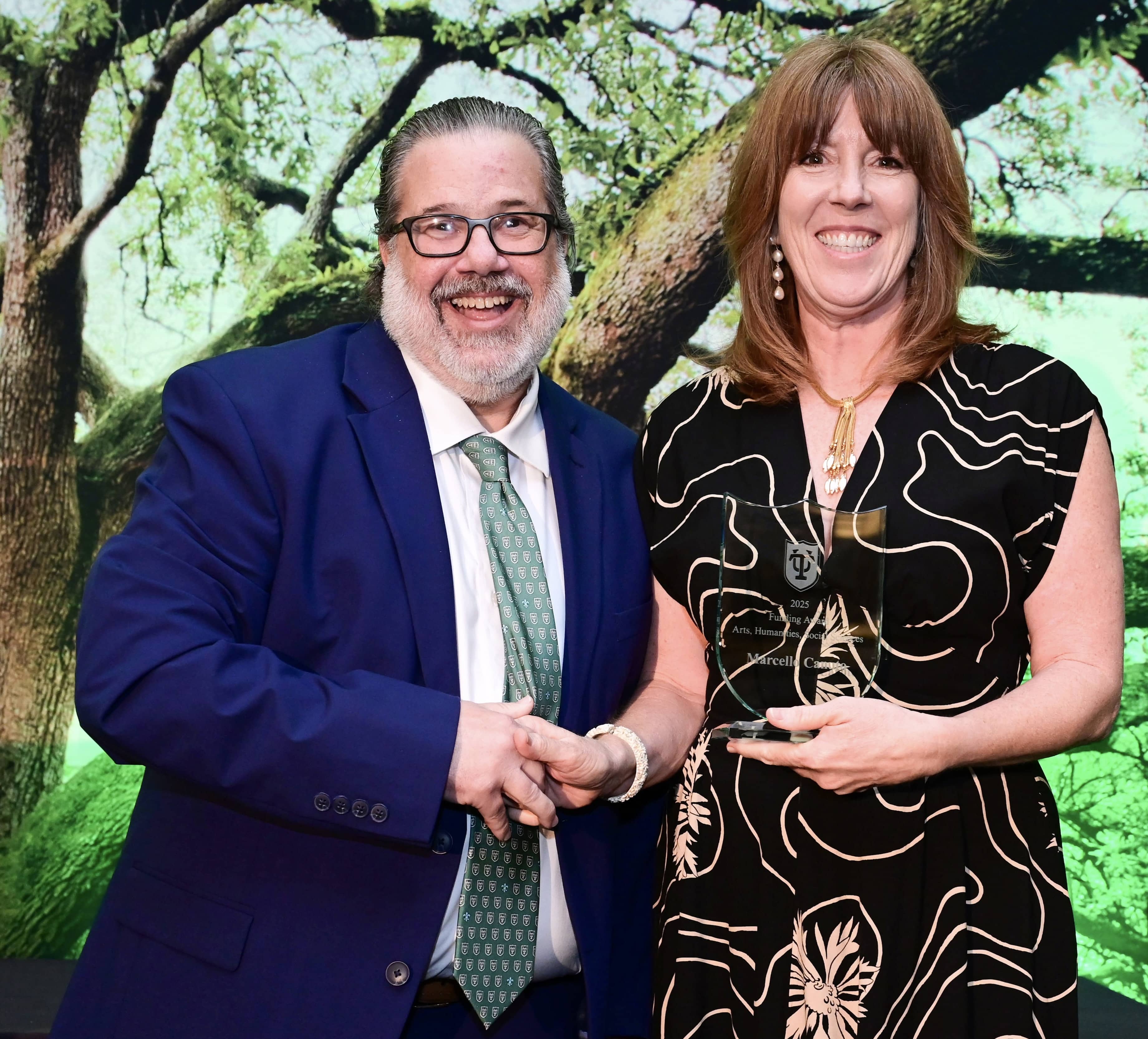 Smiling man and woman hold a glass award against a green tree-patterned backdrop.