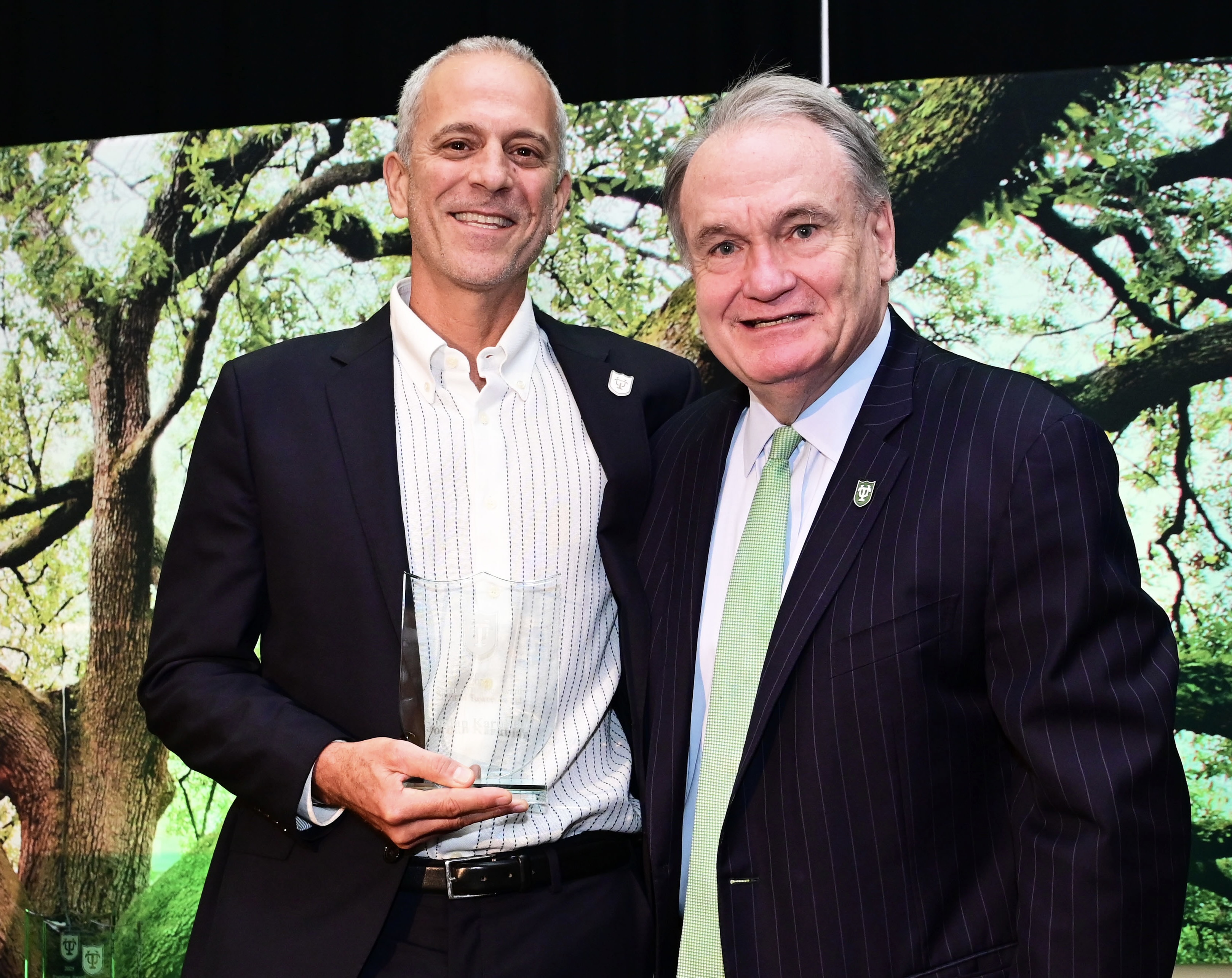 Two smiling men pose, one holding a glass award, against a green tree graphic.