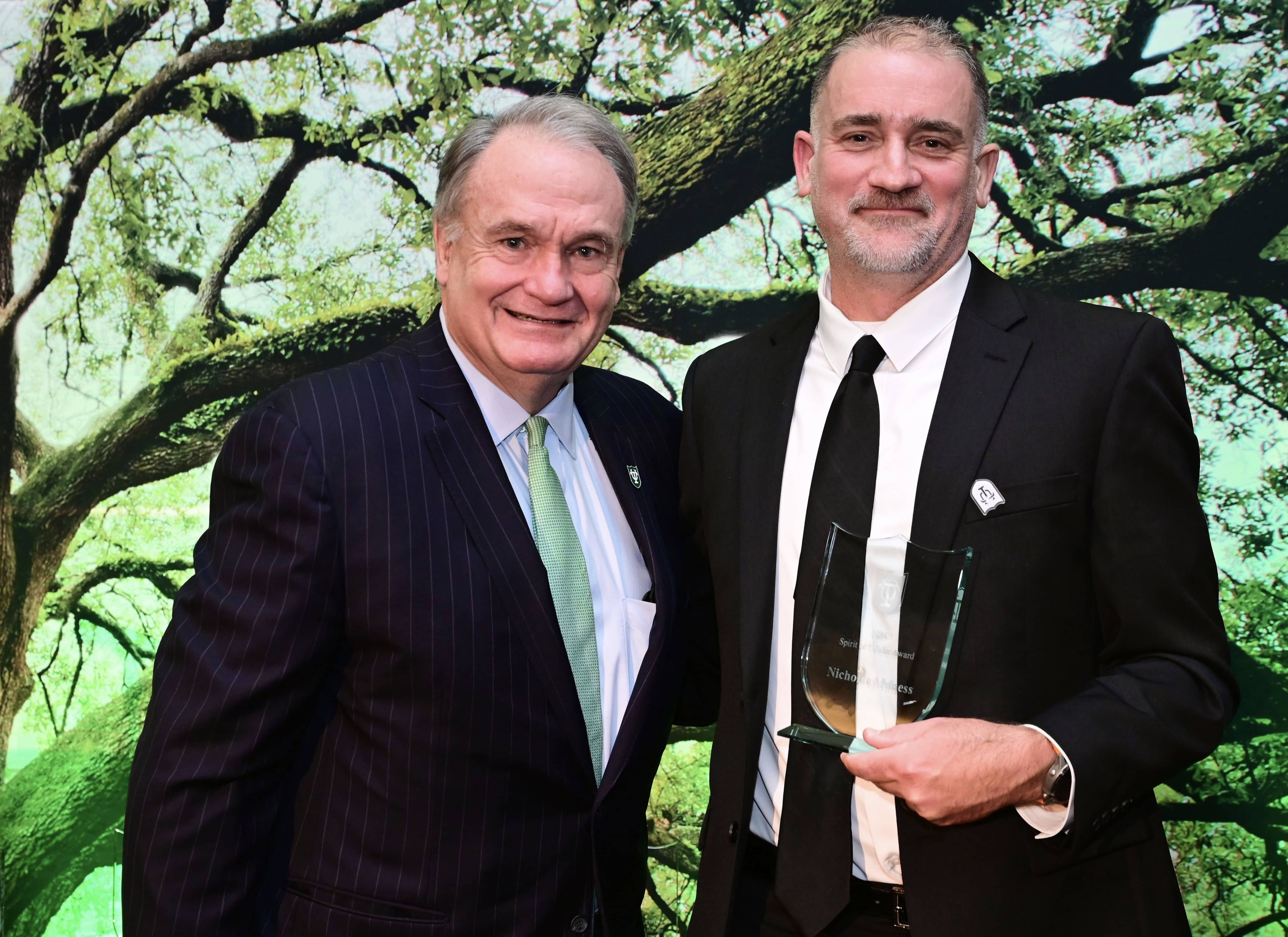 Two smiling men in suits, one holding a glass award, against a green tree backdrop.
