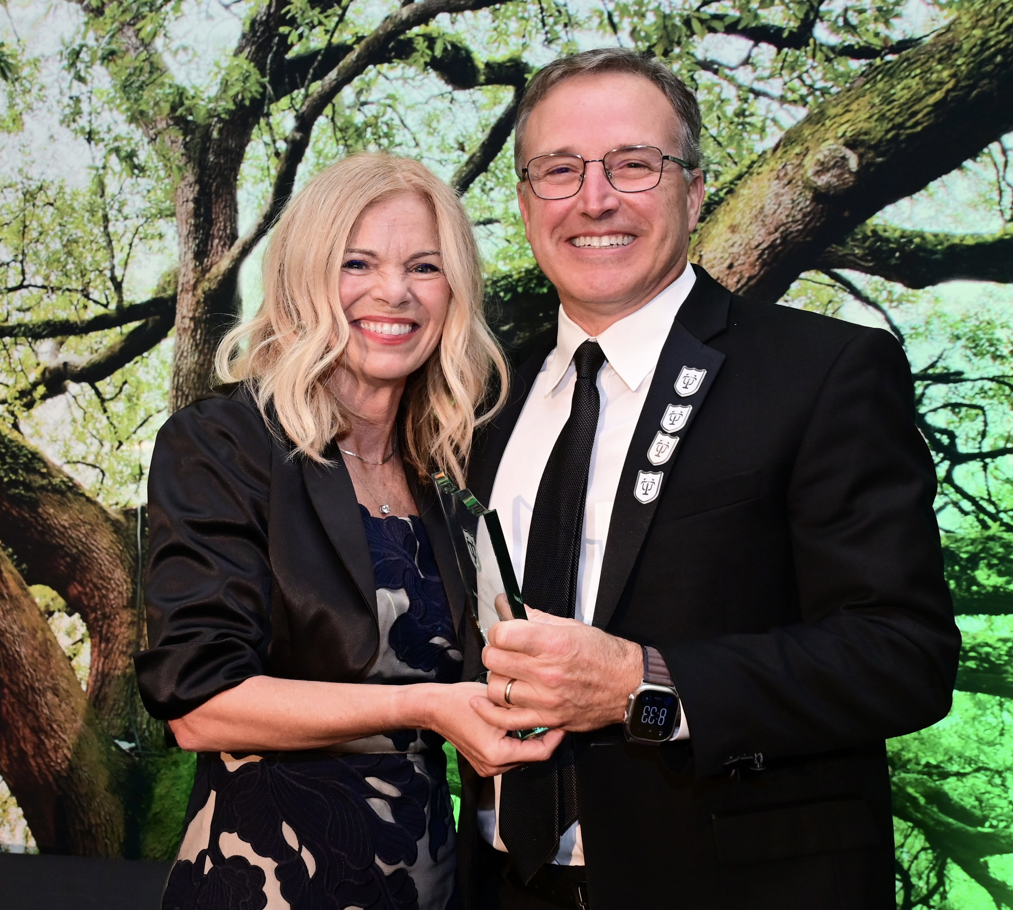 Man and woman smiling, jointly holding a green award trophy. Large tree backdrop.