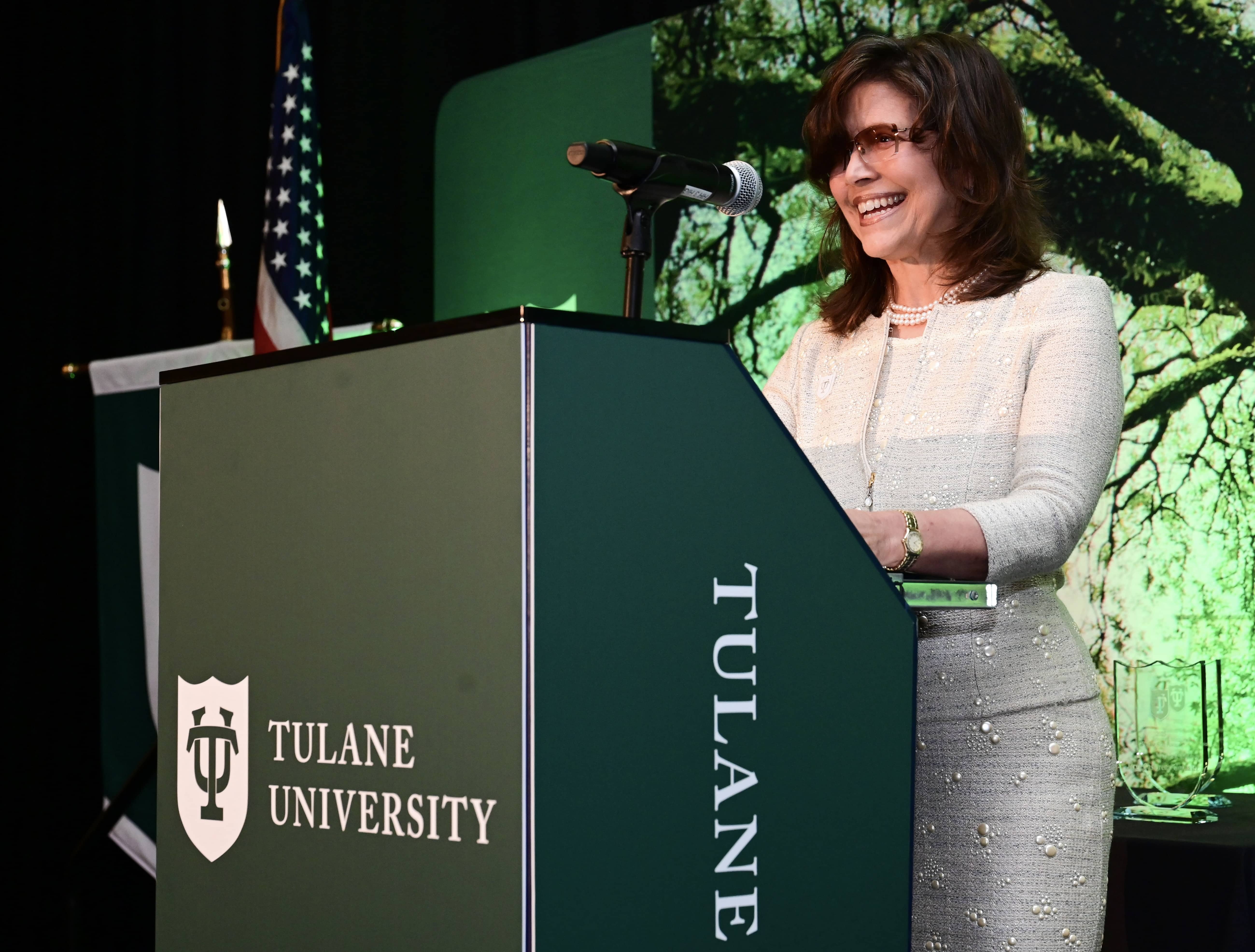 Smiling woman in glasses speaks at Tulane University podium with American flag.