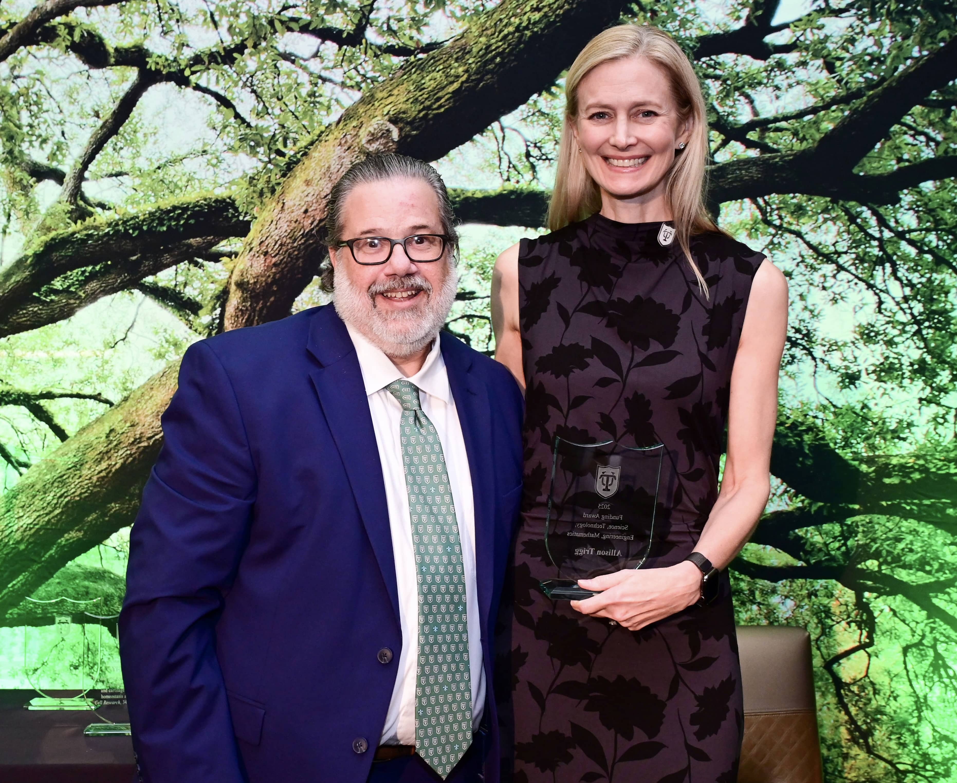 Man in blue suit and woman in dark dress holding an award, smiling against a tree backdrop.