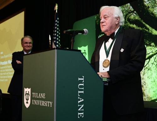 Man in tuxedo with medal speaks at green Tulane University podium; another man smiles behind.