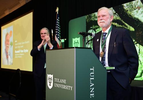 Man with medal stands at Tulane podium; another man applauds on stage.